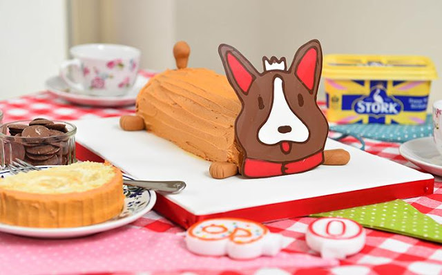 A Corgi Cake shaped like a cartoon pup, with icing details and a playful face, sits on a white board. Plates, cups, candles, and a tub of Stork margarine surround it on a red-checked tablecloth. Perfect for any Corgi Cake Recipe!.