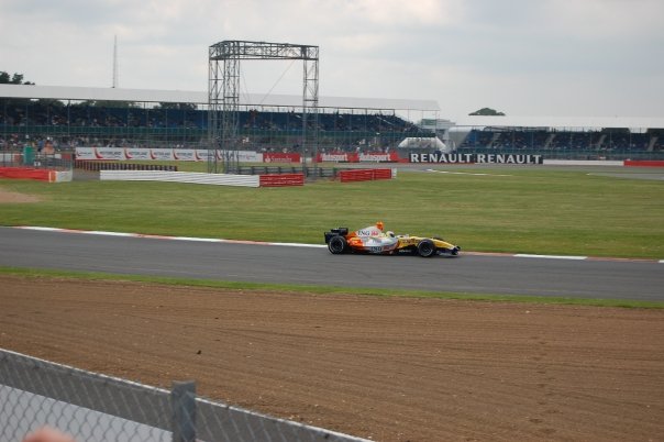 A yellow and orange Formula 1 car races on a track, passing empty grandstands and Renault banners on a cloudy day—a perfect Flashback Friday moment for motor racing fans.