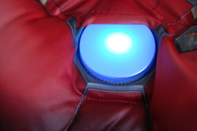 Close-up of a glowing blue circular light embedded in the chest of a red Iron Man costume, resembling the iconic arc reactor from Marvel comics and films.