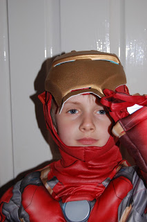 A child wearing an Iron Man costume lifts the helmet’s visor slightly with one hand and looks at the camera. This playful Iron Man costume review is set against a simple background of a white door.