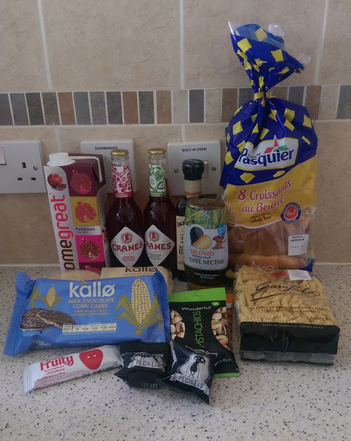 A selection of groceries from the May Degustabox subscription box on a kitchen worktop, including croissants, cranberry juice, chocolate corn cakes, mixed nuts, date nectar, fruity bars, oatcakes, Omega juice, and snack bars.