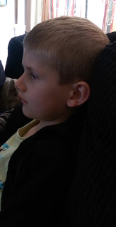 A young boy with short blond hair sits on a dark sofa, looking intently to the left, reminiscent of Charlie Brown. Bright natural light streams from a window in the background.