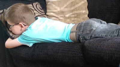 A young boy wearing glasses, a light blue shirt, and jeans is lying face down on a dark-coloured sofa with patterned cushions, reminiscent of Charlie Brown after a tough day.