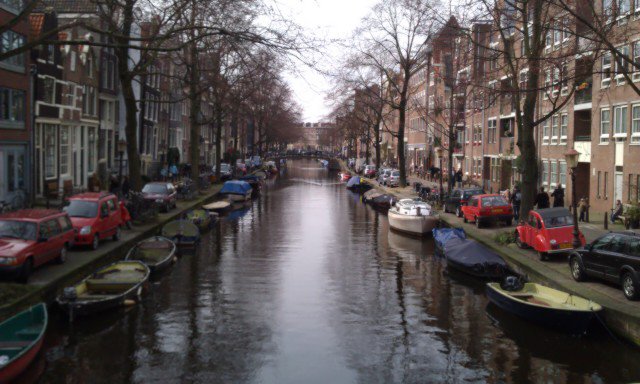 Flashback Friday: A canal in a European city lined with parked boats and cars on both sides, bordered by tall, narrow brick buildings and leafless trees on a cloudy day.