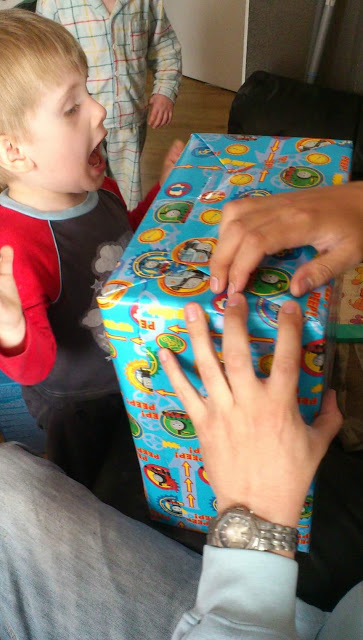 A young child with an excited expression watches as an adult’s hands begin to unwrap a large, brightly coloured present covered in cartoon character wrapping paper—an unforgettable flashback Friday moment. Another child stands in the background.