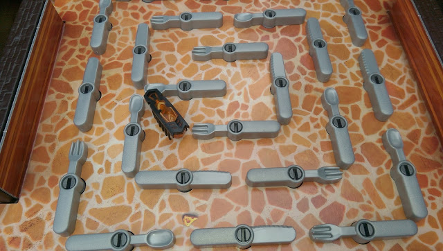 A small black and orange robotic toy scurries through a maze of grey plastic obstacles in the Bugs in the Kitchen game, navigating twisting paths blocked by adjustable levers on a board designed to look like stone tiles.