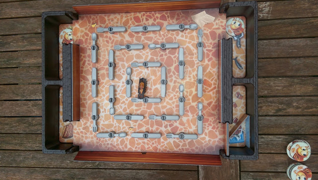 A board game with a maze pattern, numbered squares, and several grey barriers—reminiscent of the Bugs in the Kitchen game. Small round animal counters are at the corners and sides, and the board rests on a wooden surface.