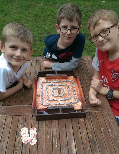 Three young boys are sitting at a wooden outdoor table, smiling at the camera as they play the Bugs in the Kitchen game with its square board and counters. Grass is visible in the background.