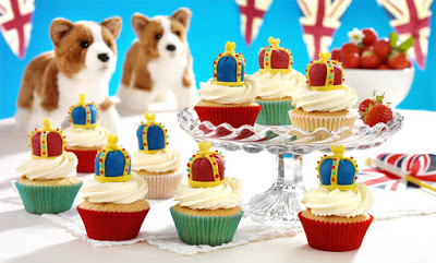 Cupcakes with royal crown toppers are displayed on a glass stand and table, alongside strawberry cream cakes, toy corgis, a bowl of strawberries, and British flag bunting in the background.
