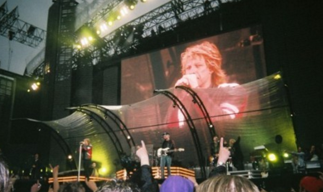 A rock band performs on a large, brightly lit stage at an outdoor concert for Flashback Friday, with a giant screen behind them displaying a close-up of the lead singer holding a microphone. Members of the audience are visible in the foreground.