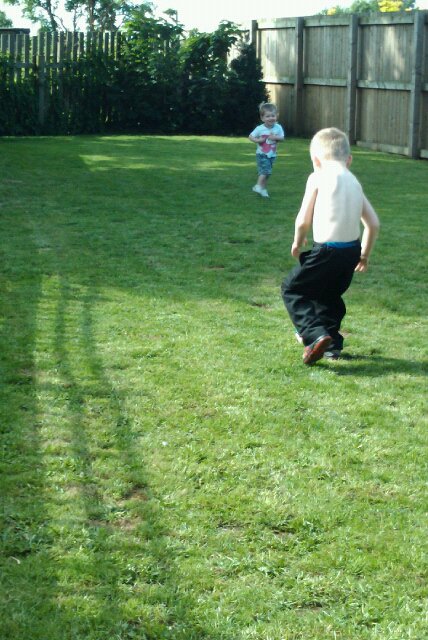 A young boy wearing black trousers and no shirt runs on grass towards a toddler in a garden, with wooden fences and bushes in the background—a classic parenting moment lit by summer sunshine, with shadows stretching across the lawn.