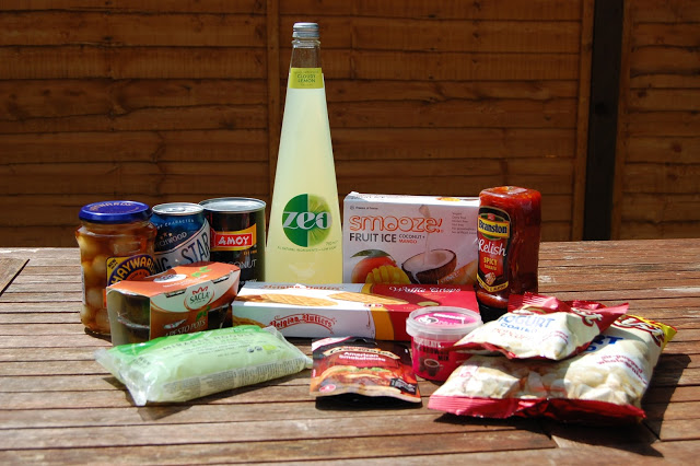 A variety of packaged foods and drinks, including jars, tins, fruit ice, a bottle of Zeo drink, snacks, and preserves from a Degustabox are displayed on a wooden table outdoors with a wooden fence in the background.