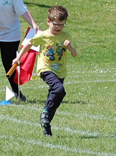 A young boy wearing glasses, a yellow T-shirt, and dark trousers runs on a grassy field holding a baton, participating in a race. Flashback Friday to those fun childhood sports days—another person holding flags is visible in the background.