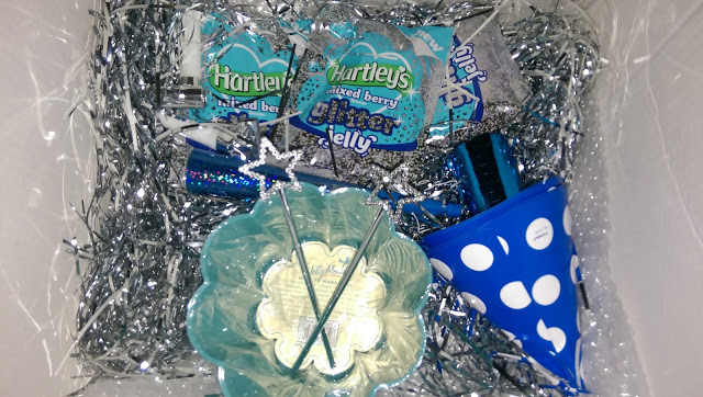 A gift box filled with silver tinsel, a blue polka dot party hat, blue bowl, silver glittery star wands, noise maker, and two packets of Hartley’s Mixed Berry Glitter Jelly—perfect for your own Harleys Glitter Jelly Review party!.