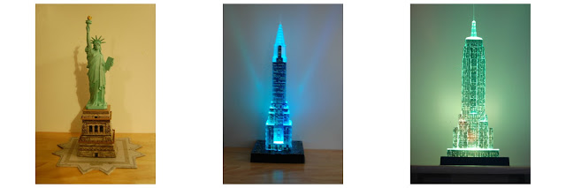 Review three Ravensburger 3D Puzzles featuring famous New York City buildings: the Statue of Liberty, a blue-lit Empire State Building, and a green-lit Empire State Building, each showcased against plain backgrounds.