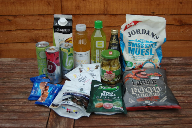 A selection of packaged snacks and drinks from a snack subscription box—including muesli, crisps, crackers, fruit infusions, energy drinks, juice, tea, bars, and a jar of pickles—displayed on a wooden table against a wooden fence.