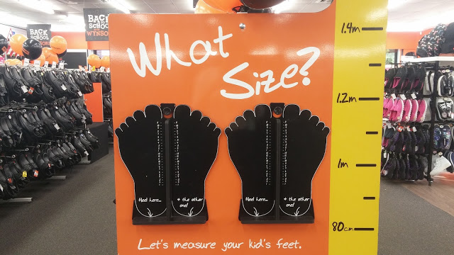 A Wynsors shoes display features two large black foot measuring charts on an orange board labelled "What Size?" and a yellow height chart on the right, with shelves of shoes visible in the background.