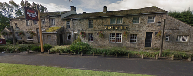 A long stone building with a sign reading “Anchor Inn” on the wall, surrounded by greenery and flowers, with a Brewer’s Fayre sign visible on the left side of the road, inviting guests to enjoy classic pub food.