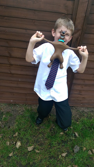 A young boy in a school uniform and striped tie stands outside on grass, playfully stretching a Stretch Scooby Doo toy across his face, squinting and smiling in front of a wooden fence.