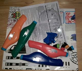 Three colourful plastic toy bottles and blue toy parts, likely from an IDO3D Vertical set, are scattered on printed instruction sheets and a black-and-white patterned surface on a wooden floor—perfect for an IDO3D Vertical review.