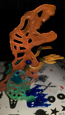 A colourful, abstract dinosaur skeleton made with a 3D drawing pen stands on a white surface decorated with black icons and shapes. The sculpture features orange, green, blue, and yellow hues with an open, skeletal design.