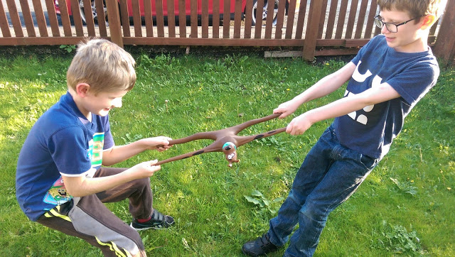 Two young boys playfully pull on a Stretch Scooby Doo toy outdoors on green grass, each holding an end and smiling. A wooden fence and parked cars are visible in the background—perfect for a fun review.