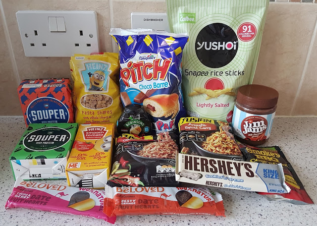 A selection of assorted snacks and convenience foods from a snack box, including instant noodles, soup cartons, rice sticks, date bars, chocolate bars, nut butter, and packaged baked goods, arranged on a kitchen worktop.