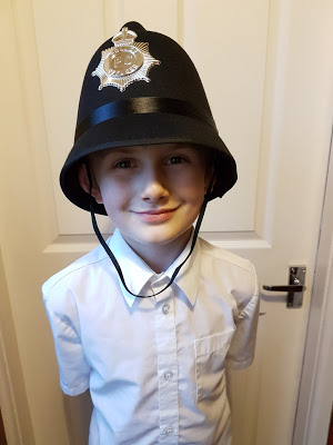 A young child wearing a white shirt and a British police officer’s helmet stands smiling in front of a closed door indoors, ready to fuel your dreams with imagination.