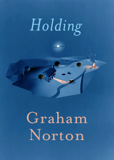 Book cover for "Holding" by Graham Norton, perfect for your September or October reading list. It features an illustration of a house with a lit path in a rural landscape at night under a glowing moon, all in shades of blue.