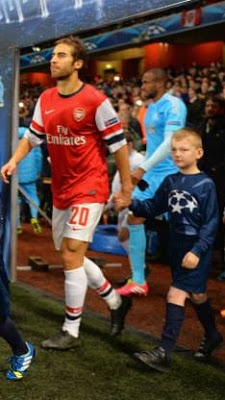 A footballer in an Arsenal shirt walks onto the pitch holding hands with a young mascot, inspiring others to fuel your dreams, while other players and children are seen in the background.