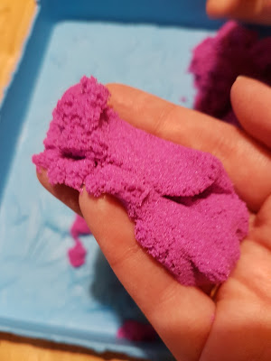 A hand holds a lump of bright purple Kinetic Sand above a blue tray, with more sand in the background—perfect for a sensory toy experience and an ideal highlight for any Kinetic Sand review.