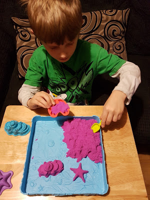 A young child in a green shirt enjoys the sensory toy, Kinetic Sand, on a blue tray, using plastic moulds and tools to create shapes like a star and seashells on a wooden table.