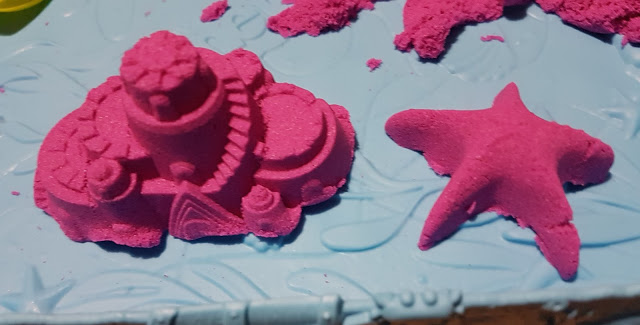 Two pink Kinetic Sand sculptures, one shaped like a castle and the other like a starfish, sit on a light blue textured surface—perfect as a sensory toy for creative play.