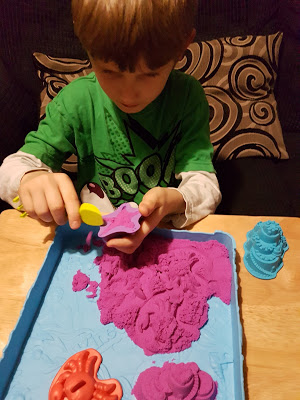 A young child wearing a green shirt plays with purple Kinetic Sand on a blue tray, using a yellow tool and moulds to shape the sensory toy.