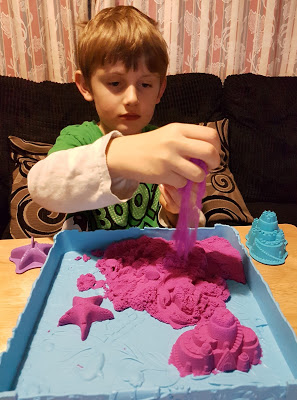 A young boy enjoys sensory play with purple Kinetic Sand in a blue tray, shaping it into starfish and castle moulds on a table indoors.
