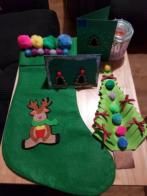 A green felt Christmas stocking with a reindeer design is displayed on a table alongside handmade cards, a paper tree decorated with pom-poms, a glass jar, and more colourful pom-poms—perfect inspiration for Christmas decorations you can Make It Don’t Buy It.
