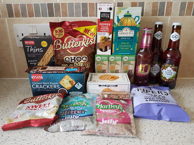 A selection of snacks and drinks from a Christmas Degustabox is displayed on a kitchen worktop, featuring biscuits, popcorn, jelly, oat biscuits, crisps, tea, sauce, and two bottles of cider—all arranged in rows against a tiled splashback.