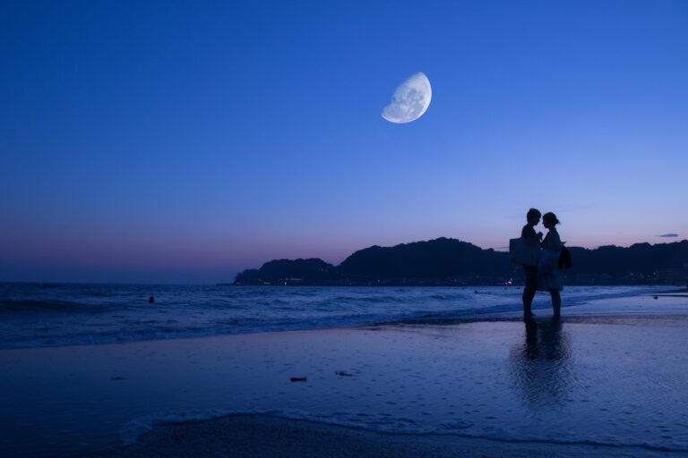A couple embraces on a beach at dusk, silhouetted against a deep blue sky with a large half moon—a perfect scene for romantic date ideas, as distant hills and moonlight are reflected in the calm water.