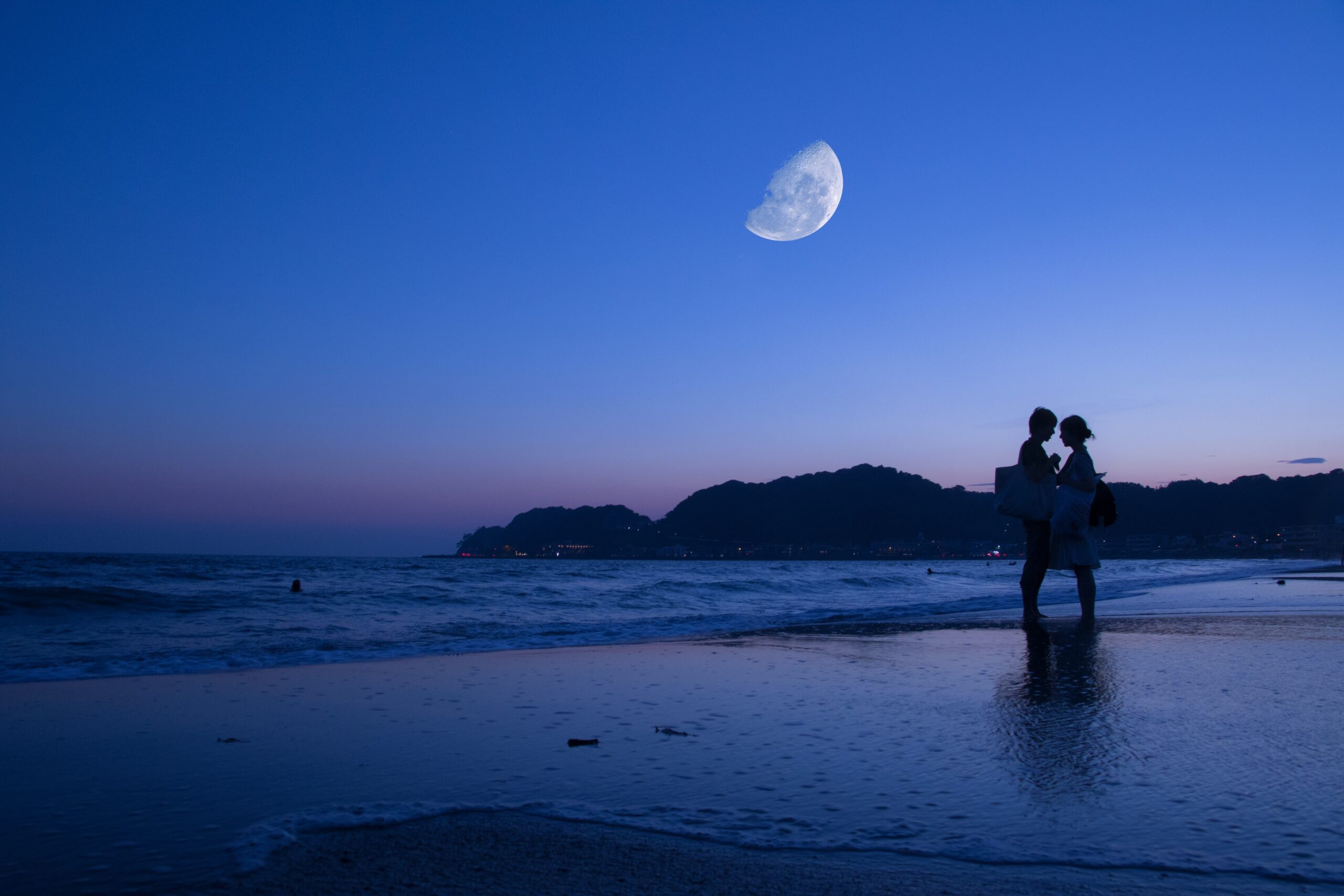 A couple embraces on a beach at dusk, silhouetted against a deep blue sky with a large half moon—a perfect scene for romantic date ideas, as distant hills and moonlight are reflected in the calm water.