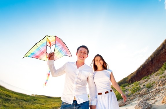 A smiling couple in white clothes stands outdoors on a grassy hill, holding hands—a perfect scene for Romantic Date Ideas. The man holds a colourful rainbow-striped kite beneath the clear blue sky, with rocks and slopes in the background.