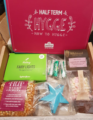 A gift box labelled "Half Term Hygge" brings fun and cosiness with fairy lights, a chocolate spoon, popcorn, tealight candles, mini marshmallows, and a blue star ornament, all neatly arranged for a perfect Half Term treat.
