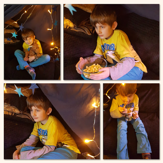 A young boy in a yellow shirt sits inside a cosy, hygge-inspired blanket fort decorated with fairy lights and paper stars, holding a soft toy and a bowl of popcorn, looking thoughtful and relaxed.
