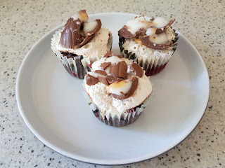 Three mini cupcakes with white icing, topped with pieces of chocolate and white drizzle, are arranged on a white plate on a light-coloured worktop—reminiscent of classic S'more Brownie Cupcakes.