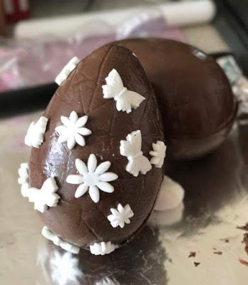 Two DIY Easter eggs decorated with white fondant shapes, including butterflies and flowers, are placed on a reflective surface.