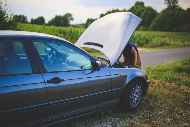 A car is parked on the roadside with its bonnet open, suggesting a car breakdown. A person inspects the engine, possibly seeking a solution, whilst greenery and fields surround the scene.