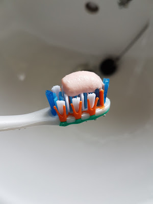 A close-up of a toothbrush with blue, white, and orange bristles holding a dab of pink toothpaste, highlighting the importance of gum health to help stop bleeding gums, with a white washbasin and drain blurred in the background.