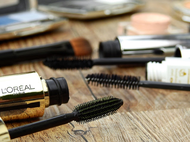 Close-up of several opened mascara tubes and brushes on a wooden surface, hinting at beauty treatments, with some compact make-up items softly blurred in the background.