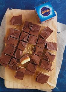 A wooden board with squares of Millionaire's Shortbread, topped with chocolate pieces, with one square partially eaten. A closed box of Cadbury Caramel sits next to the board, all set against a blue background.