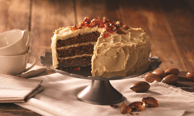 A three-layer caramel cake with light brown icing sits on a pedestal. A slice is missing, revealing moist cake layers. Pecans are scattered around the wooden table, and two stacked teacups are placed nearby.
