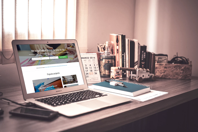 A laptop displaying an educational website sits on a wooden desk with a notebook, pen, mobile phone, desk calendar, and books—much like how a local wedding photographer might organise their workspace after booking clients through Bidvine.